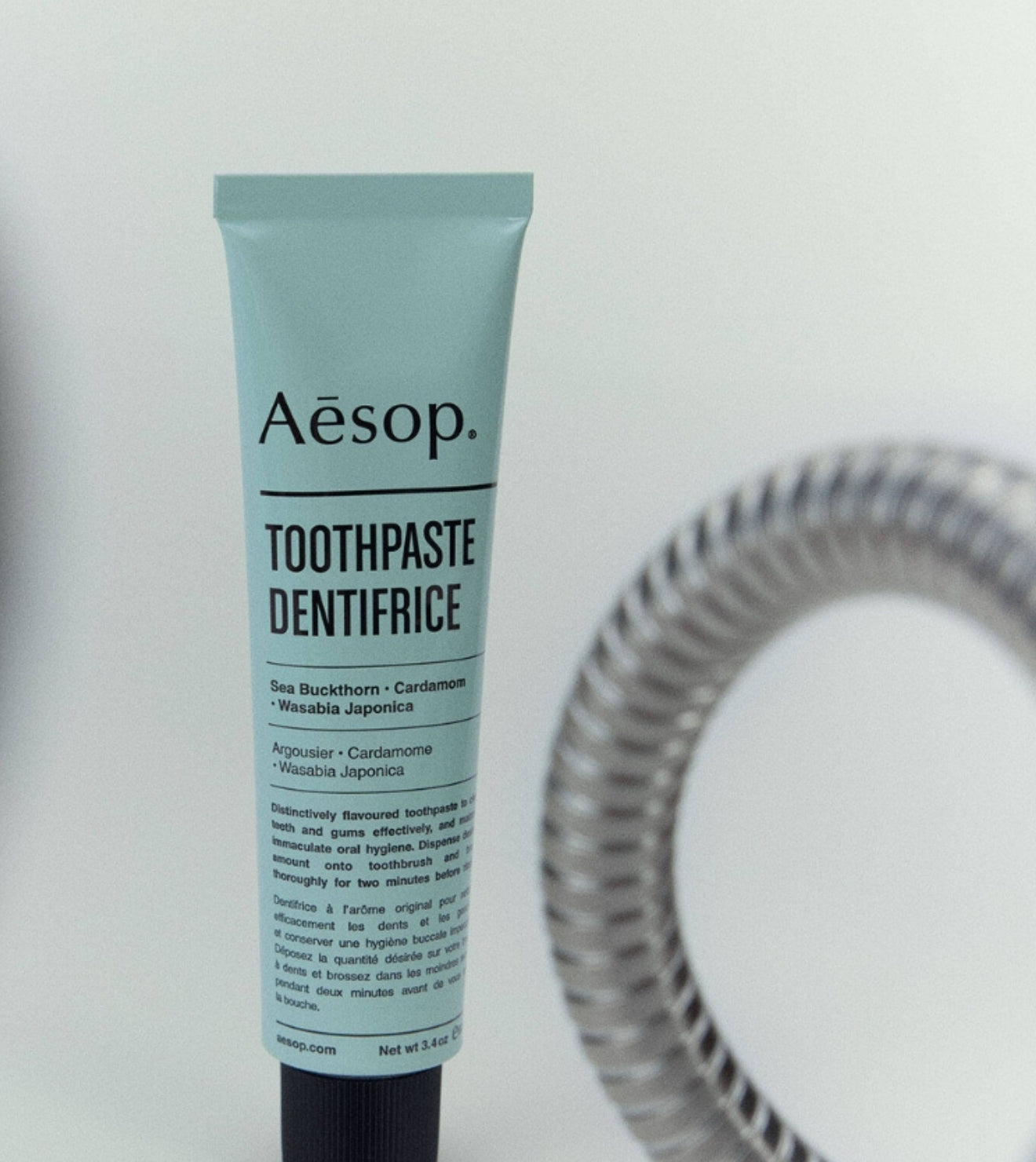 A tube of Aesop Toothpaste Dentifrice standing upright against a white background, with a blurred coiled hose in the background.