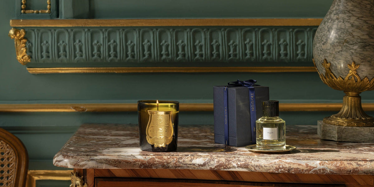 A luxury home fragrance collection displayed on a marble console table against a dark green paneled wall. The collection includes a gold shield-shaped candle in a black glass vessel, a navy blue gift box with a blue ribbon, and a small clear glass perfume bottle on a gold tray.