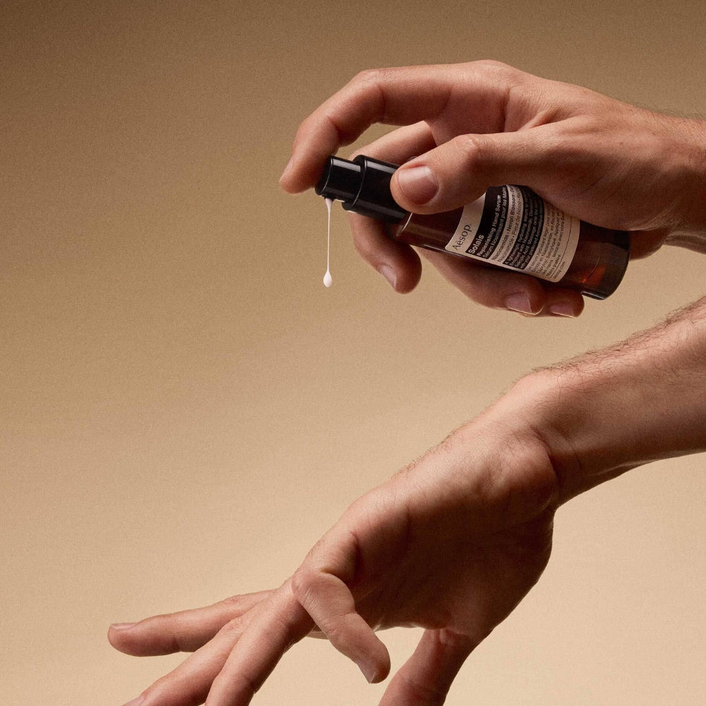 A close-up of a person's hands holding and spraying an amber glass bottle with a black pump dispenser, with a single drop of Aesop Solais Replenishing Hand Serum dripping down, against a warm beige background.