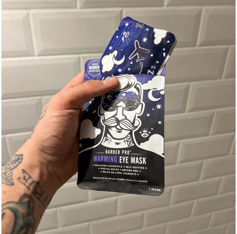 Hand holding a Barber Pro warming eye mask package against a tiled wall.