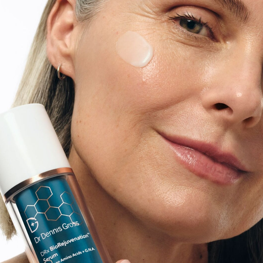 Close-up of a woman's face with a dollop of cream on her cheek, holding a bottle of Dr. Dennis Gross DRx BioRejuvenation Serum.