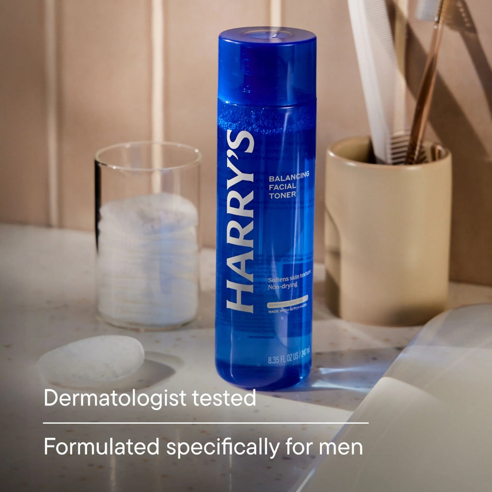 A blue bottle of Harry's Balancing Facial Toner stands on a bathroom counter with a glass of water, cotton pads, and a toothbrush holder in the background. Text at the bottom reads 'Dermatologist tested' and 'Formulated specifically for men'.