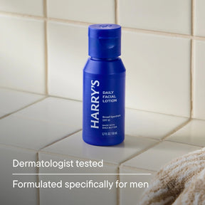 A blue bottle of Harry's Daily Facial Lotion SPF 15 stands on a beige tiled bathroom counter, with text below reading 'Dermatologist tested' and 'Formulated specifically for men'.