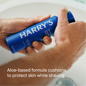Hand holding a blue 'HARRY'S' shave gel can with text about aloe-based formula.