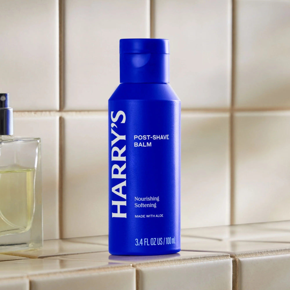 A blue bottle of Harry's Post-Shave Balm standing on a beige tiled surface, with part of a perfume bottle visible on the left.