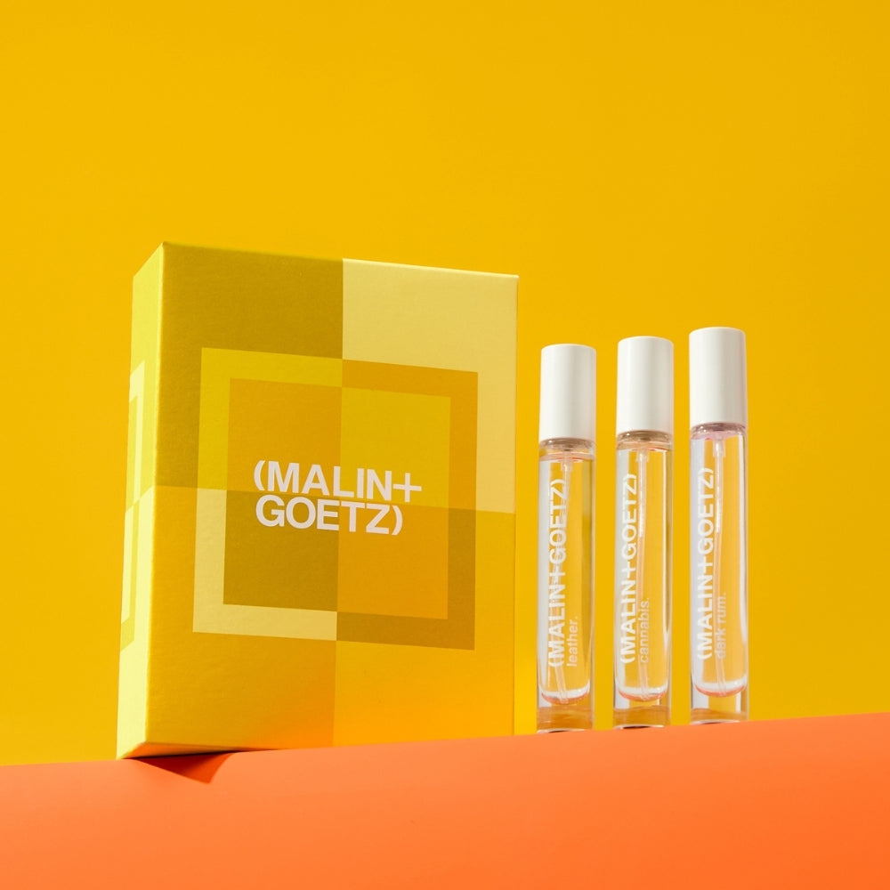 Three clear glass perfume sample vials with white caps stand beside a yellow and orange geometric box printed with the brand name (MALIN+GOETZ) on a bright yellow background.